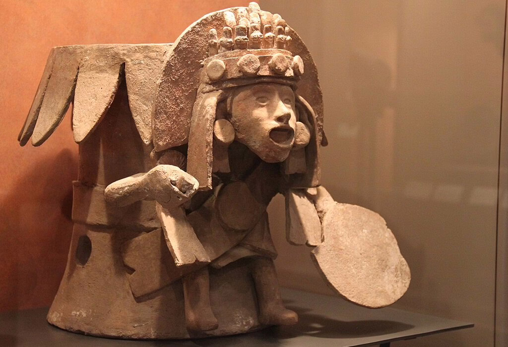 Brasero aztèque en poterie en forme de divinité avec une coiffe détaillée et un tambour, exposé dans un musée au Mexique, montrant l'art préhispanique.