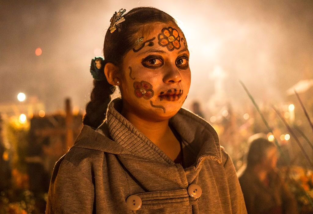 Young woman with painted face for Day of the Dead in Mixquic, Mexico, standing among candles and night celebrations.