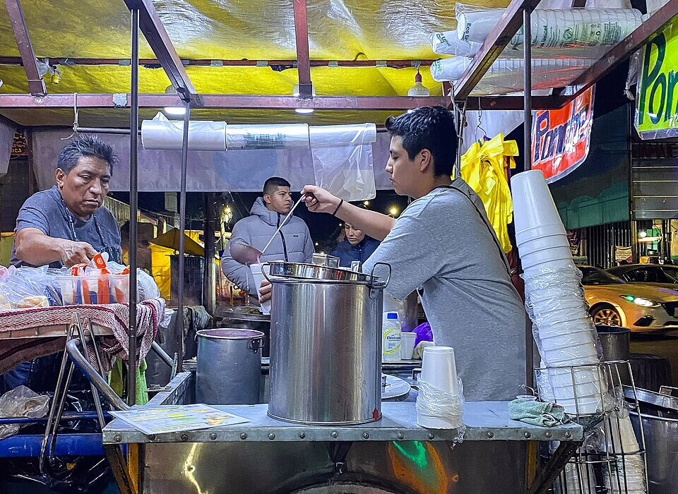 Local street food vendor serving hot tamales and atole from large metal pots at a night market in Mexico City.