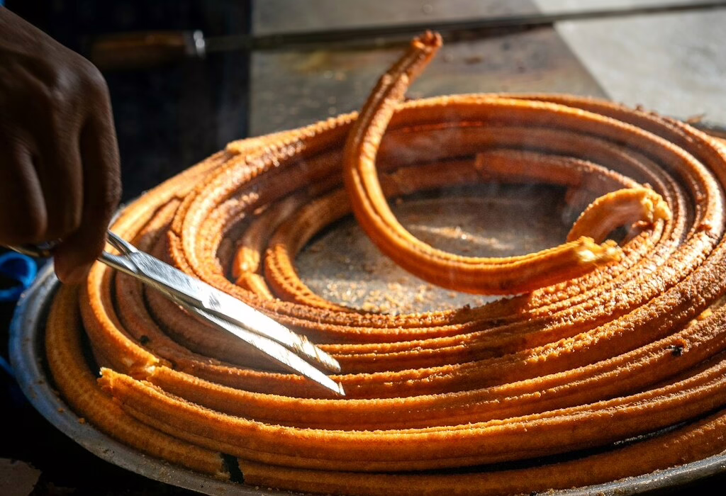 Des churros fraîchement frits en forme de spirale sont coupés à l'aide de pinces sur un étal de cuisine de rue. Ils sont dorés et prêts à être dégustés.