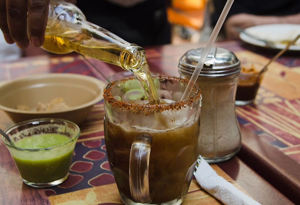 A cold Mexican michelada with beer poured into a salt-rimmed glass on a colorful table with sauces.