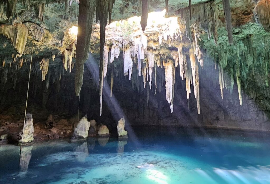 Beautiful cave with crystal-clear water and stalactites, illuminated by sunlight streaming from above.