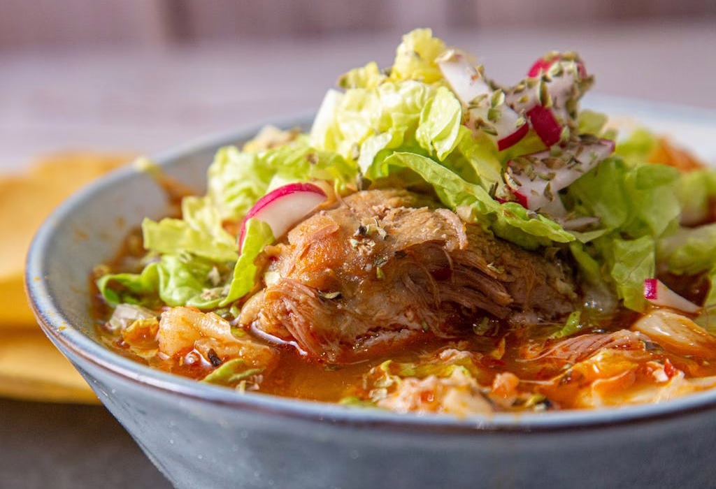 Bowl of pozole rojo with shredded pork, hominy, red broth, topped with lettuce and radish slices.