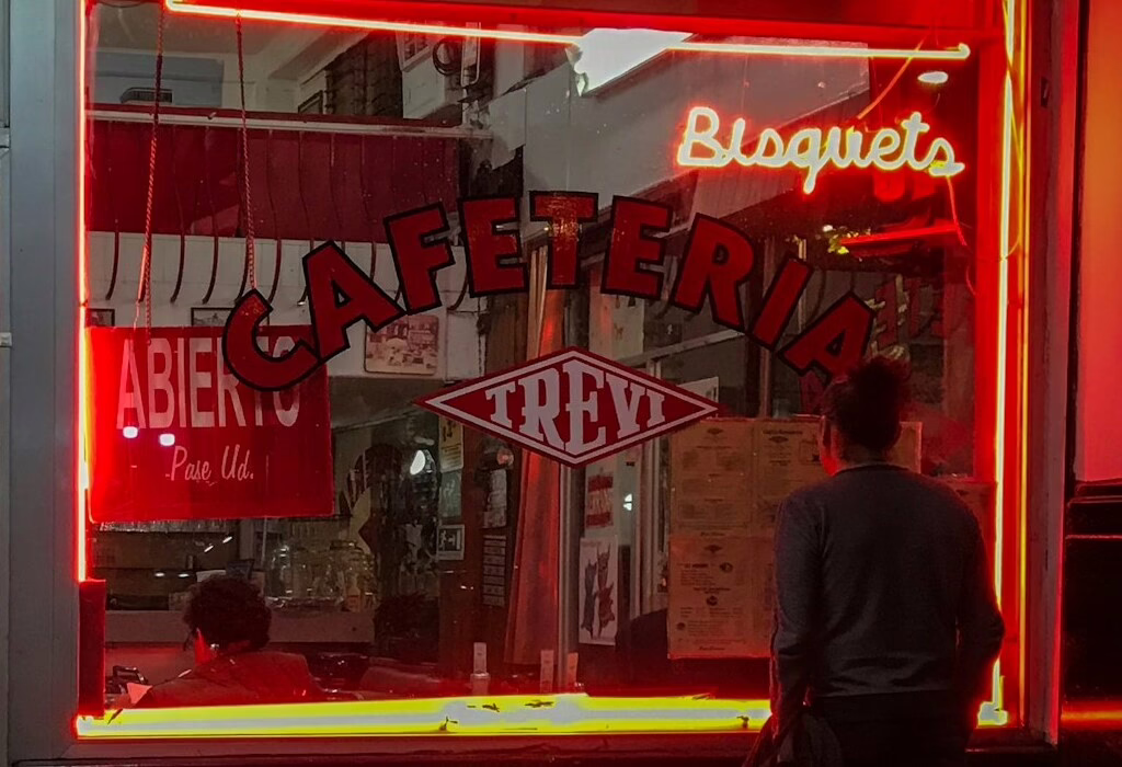Neon sign of a Mexican cafeteria glowing red at night with people inside.