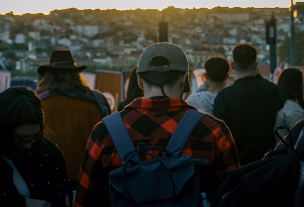 Back view of a traveler in red plaid with a backpack among a crowd at a city viewpoint during sunset, overlooking a blurred hillside cityscape.