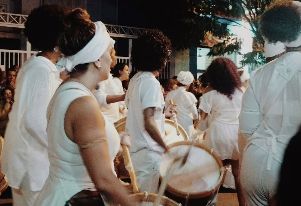 Un percussionniste en tenue traditionnelle blanche, concentre et frappe un grand tambour de cérémonie avec des baguettes lors d'une procession à faible luminosité.