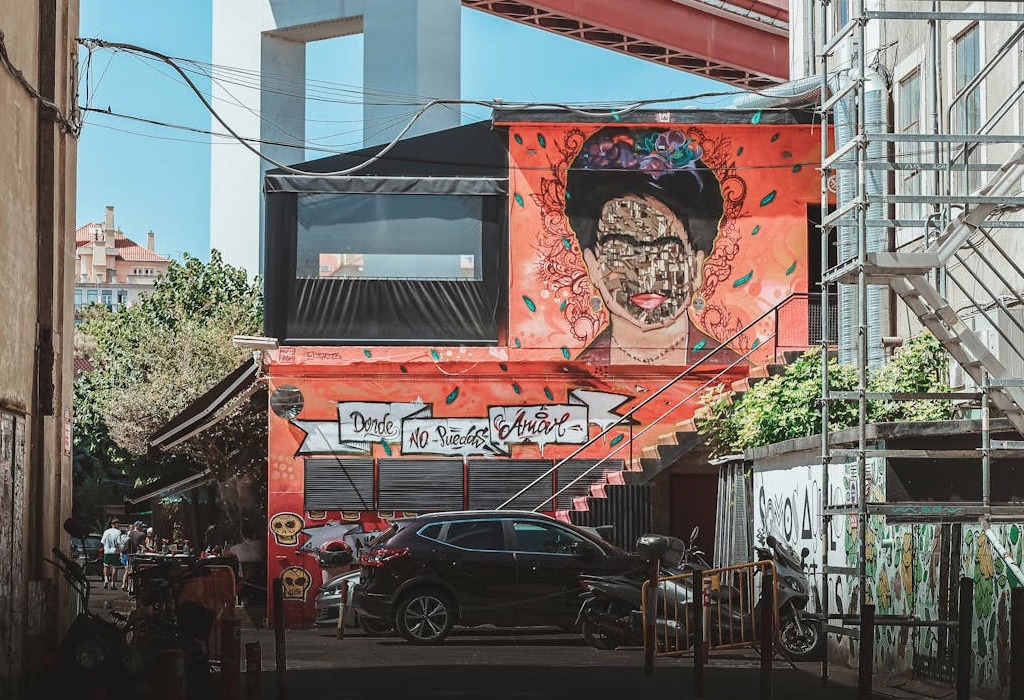 Frida Kahlo mural on an urban building in Lisbon with graffiti, cars, and a red bridge structure above.