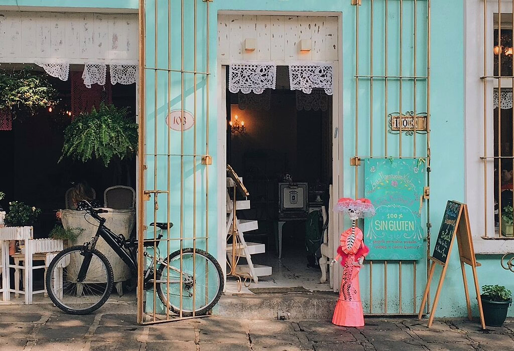 Facciata colorata di un caffè a Coyoacan, a Città del Messico, con porte aperte, piante, biciclette e una strada tranquilla, che mostra la vita quotidiana e lo stile locale amato dai viaggiatori