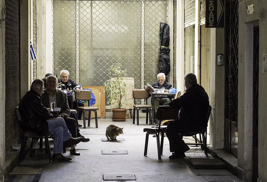 Elderly Greek men sitting and talking in a traditional kafeneio in Athens, with a cat walking in the alley.