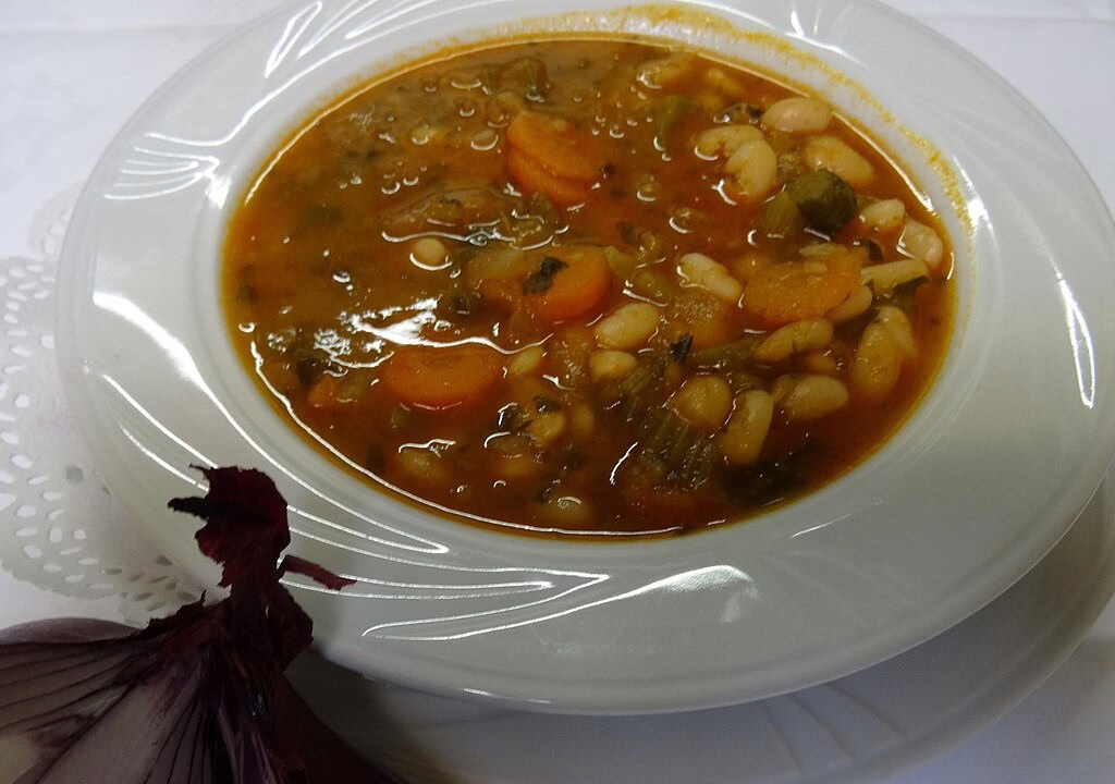 Greek fasolada soup with white beans, carrots, tomato sauce, and herbs served in a white bowl on a table.