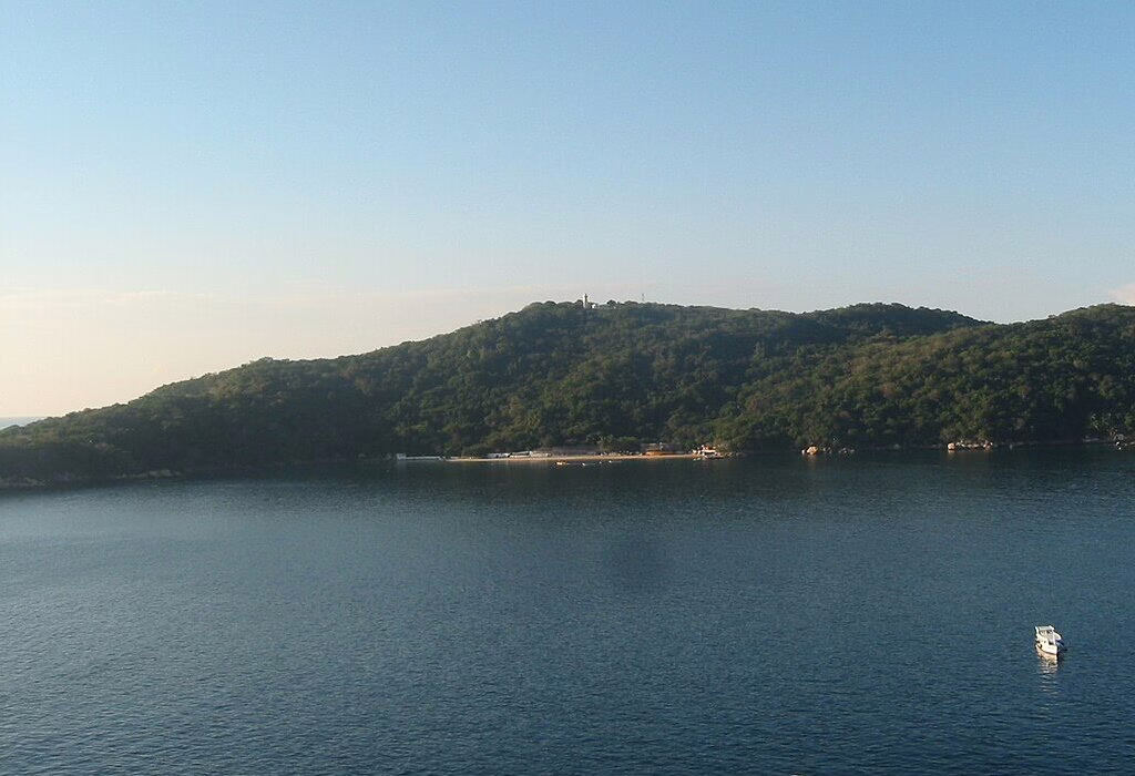 Panoramic view of the green-covered Isla Roqueta island near Acapulco, Mexico, surrounded by calm blue water and small fishing boats, a popular spot among hidden beaches in Mexico.