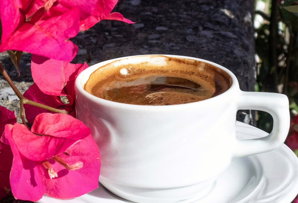 A cup of traditional Greek coffee in a white ceramic cup, placed beside bright pink bougainvillea flowers in an outdoor setting.