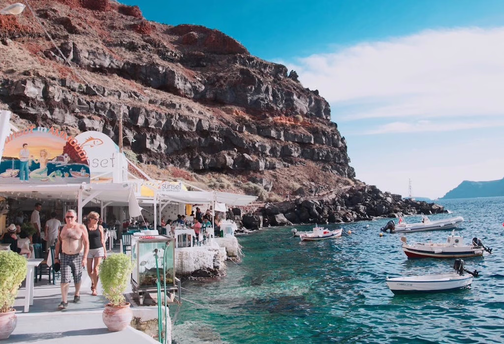 Seaside taverna in Santorini with people dining near the water, colorful boats floating, and volcanic cliffs in the background on a sunny day.