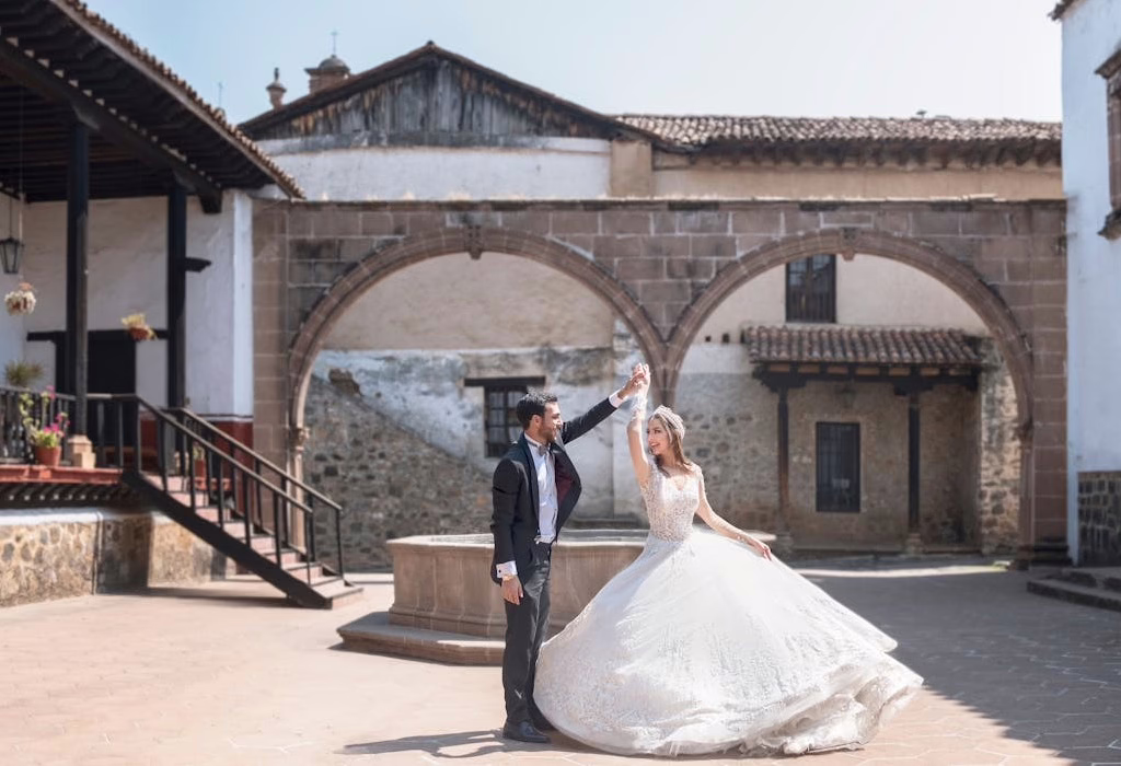 A bride in a white ballgown and a groom in a dark suit dancing in a sunlit historic Mexican courtyard with stone arches.
