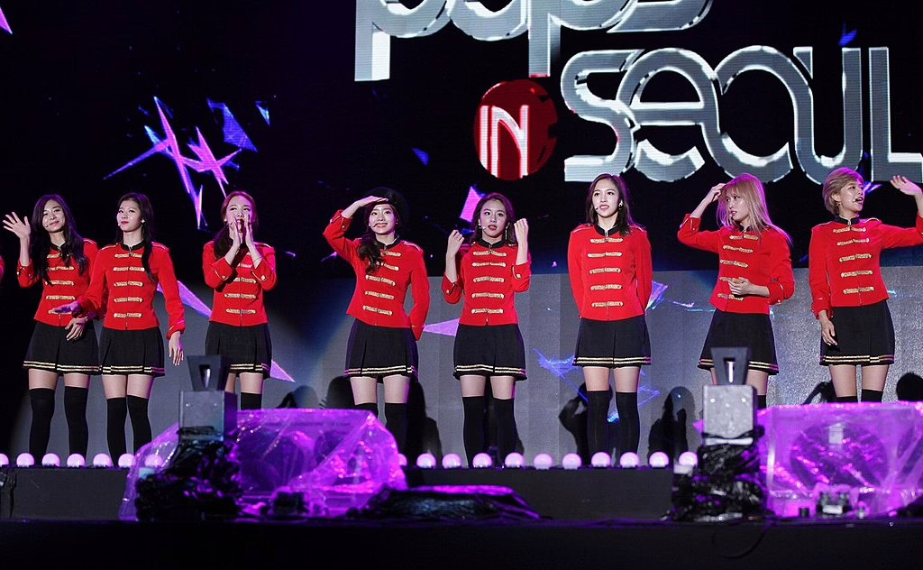 A K-pop group performing on stage in red military-style uniforms at a concert in Seoul, waving and engaging with the audience.