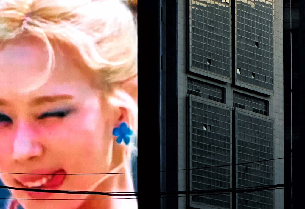 A close-up of a woman with blonde hair and a blue flower earring, with a modern building in the background.
