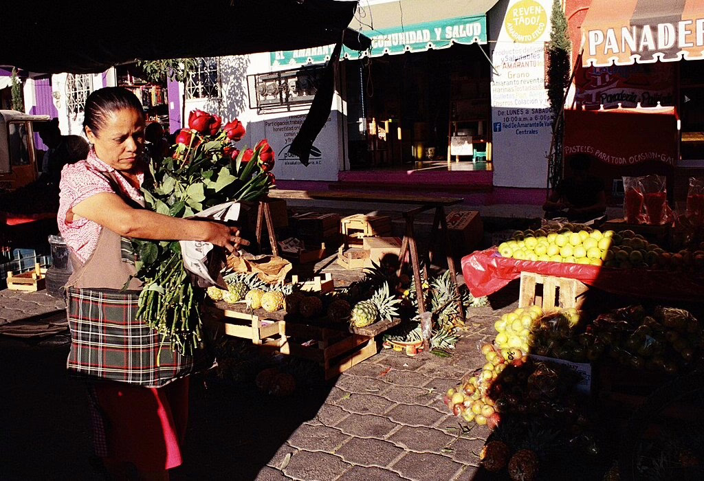 A woman carries roses at the market, surrounded by fresh fruits like pineapples and apples. A vibrant tianguis scene in Mexico.