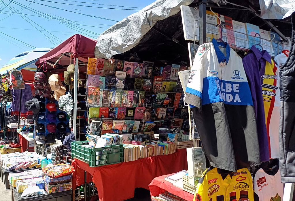 A lively street market in Mexico, featuring colorful clothing, books, and accessories for sale. The sun shines over the busy stalls in this local tianguis.