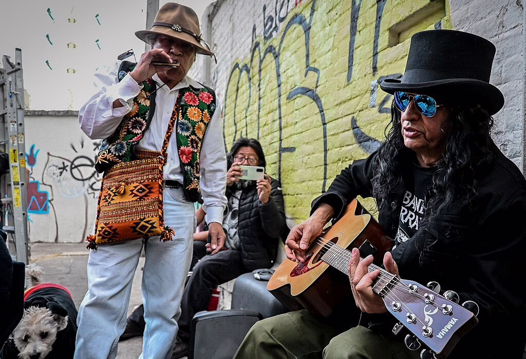 Two musicians performing at the Tianguis Cultural del Chopo, one playing a guitar and the other playing a flute, while a spectator captures the moment.