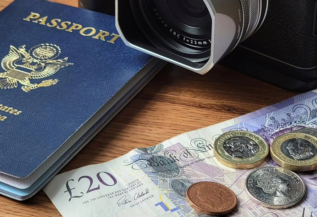 Close-up of a US passport beside a camera, with a £20 note and British coins on a wooden table, symbolizing travel planning, money exchange, and budgeting.