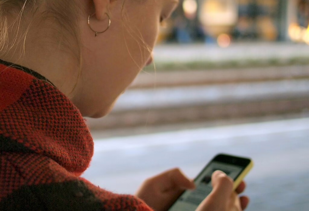 Person wearing a red checkered jacket using a smartphone while waiting near a train station platform.