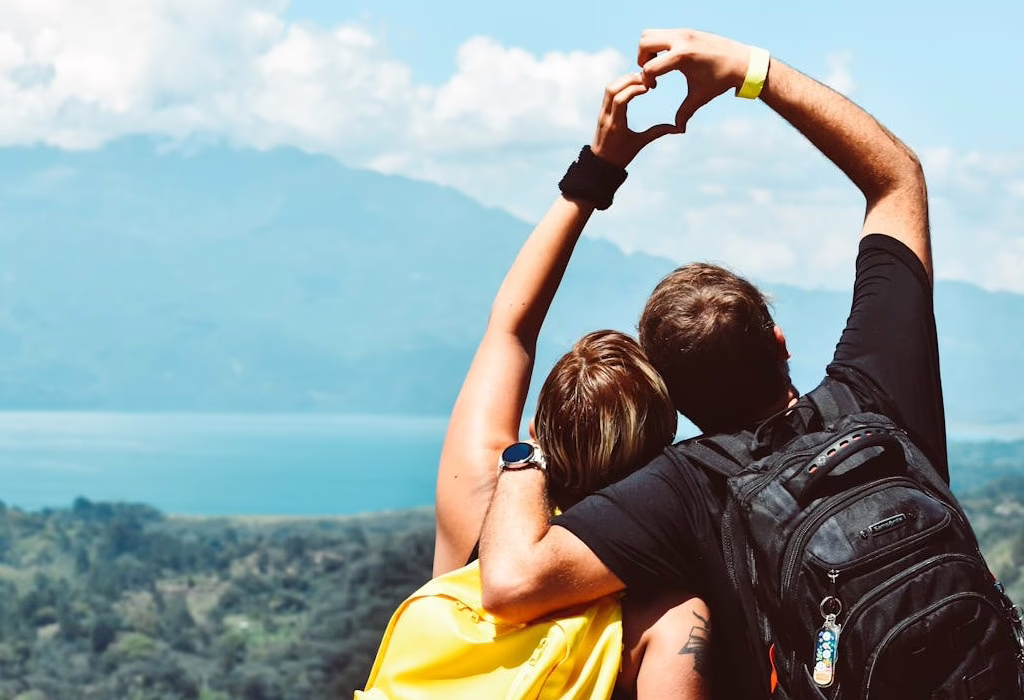Couple with backpacks making a heart shape with hands while looking at a mountain view and blue lake under a bright sky.