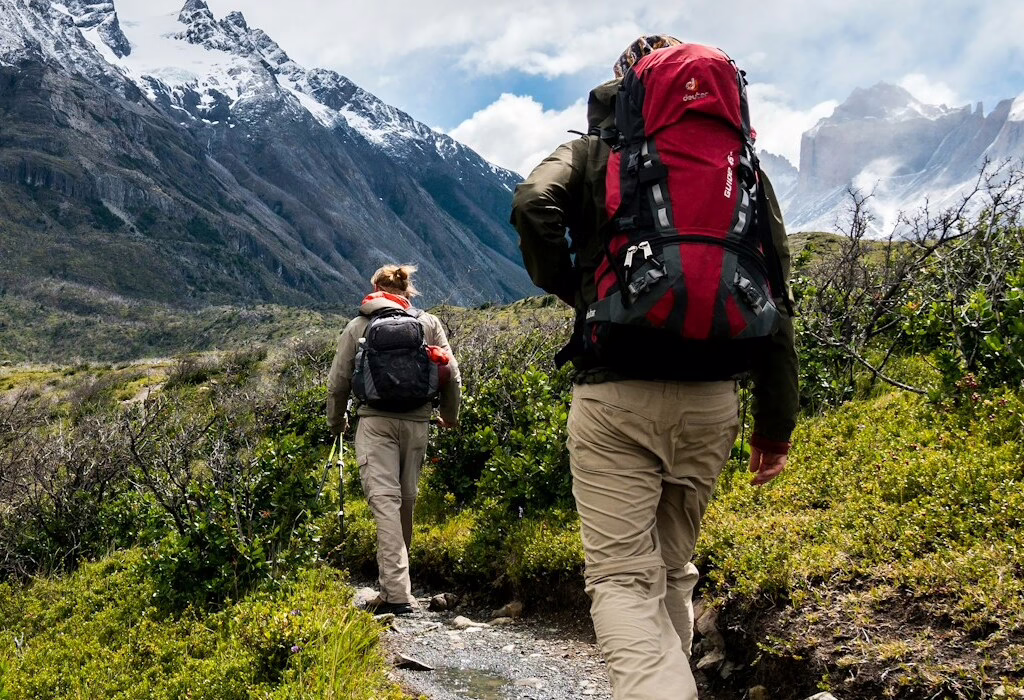 Two hikers with large backpacks walk a narrow trail through green valley toward snow-capped mountains under clouds, outdoor trekking adventure in rugged alpine scenery.