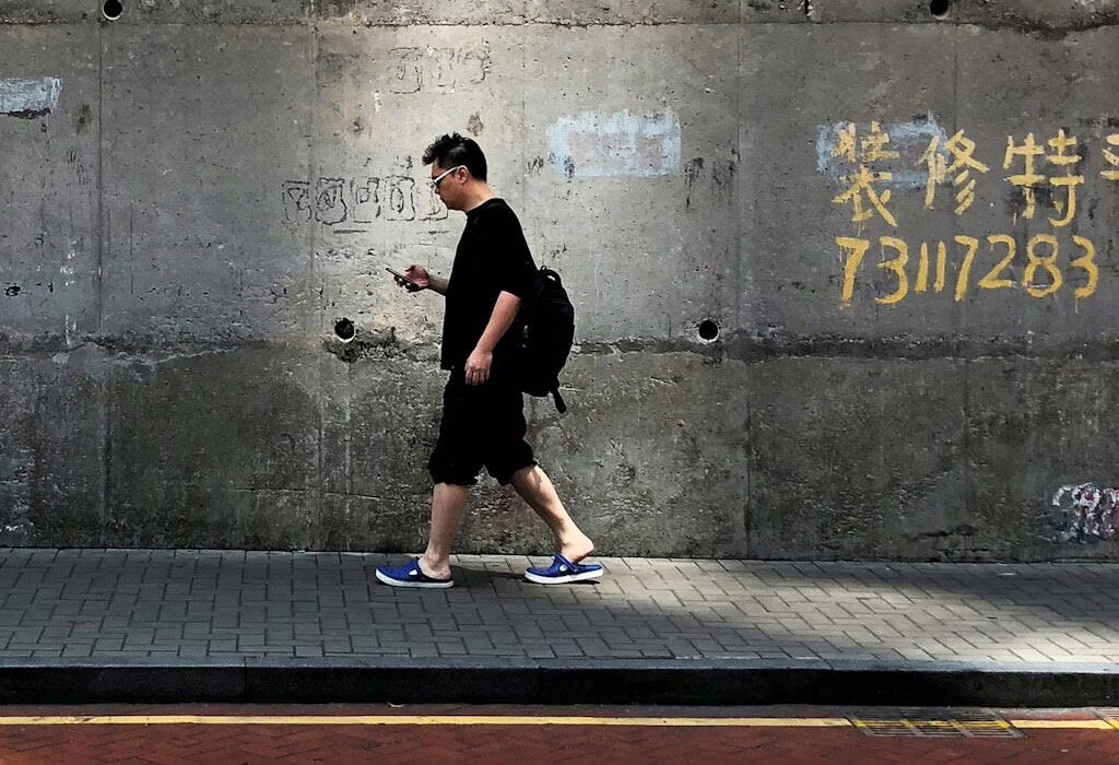 Man with backpack walking along a city sidewalk, looking at his smartphone by a concrete wall; traveler navigating streets using public wifi and maps while exploring.