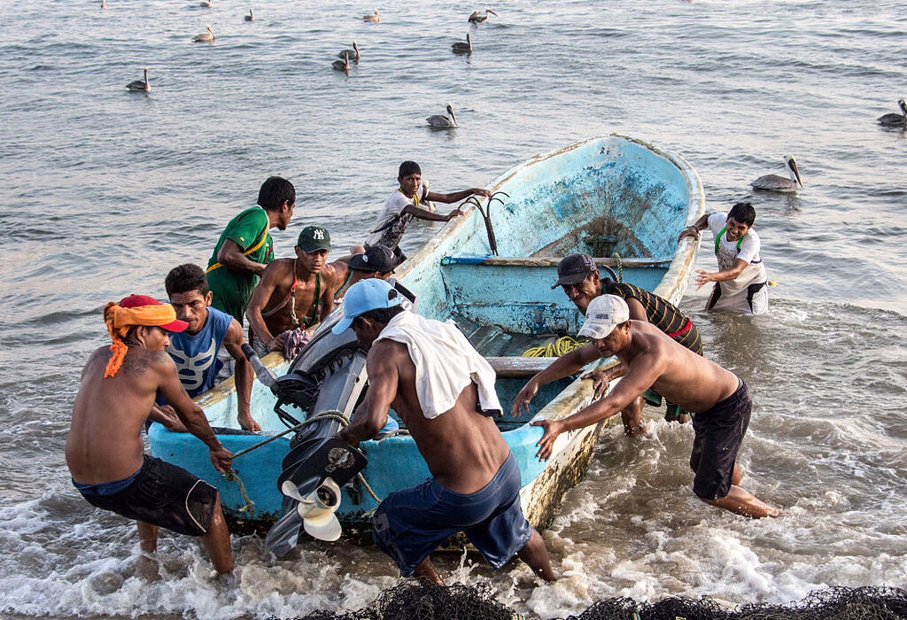 Local fishermen working together to pull a boat onto the beach in Acapulco, with the ocean and pelicans in the background.
