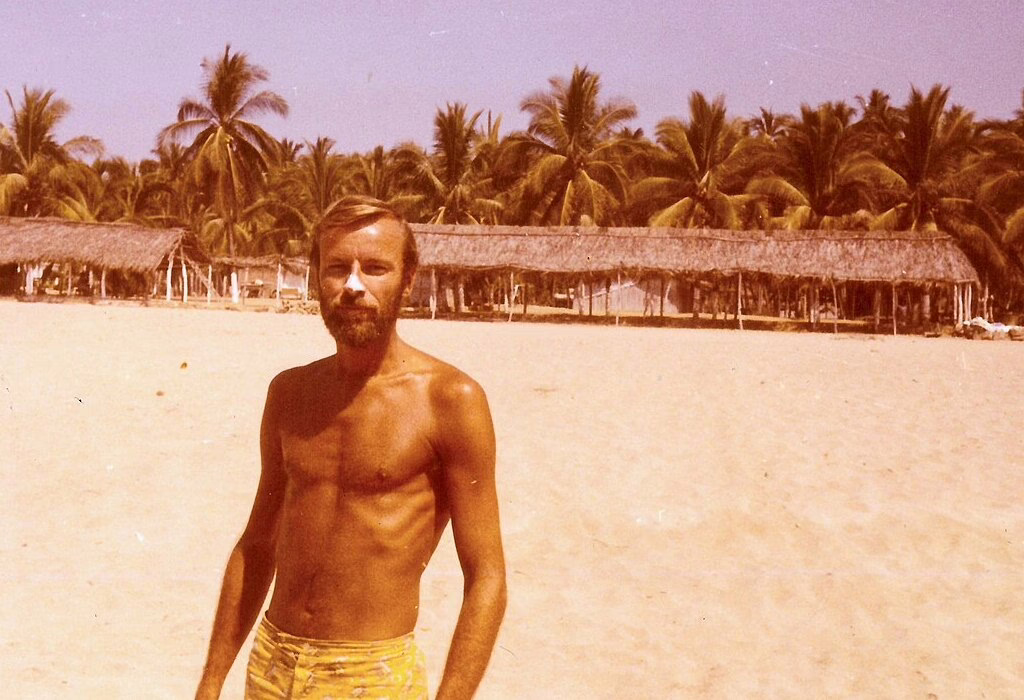 A retro photo of a man standing on the beach in Acapulco in the 70s, surrounded by palm trees and beach huts.