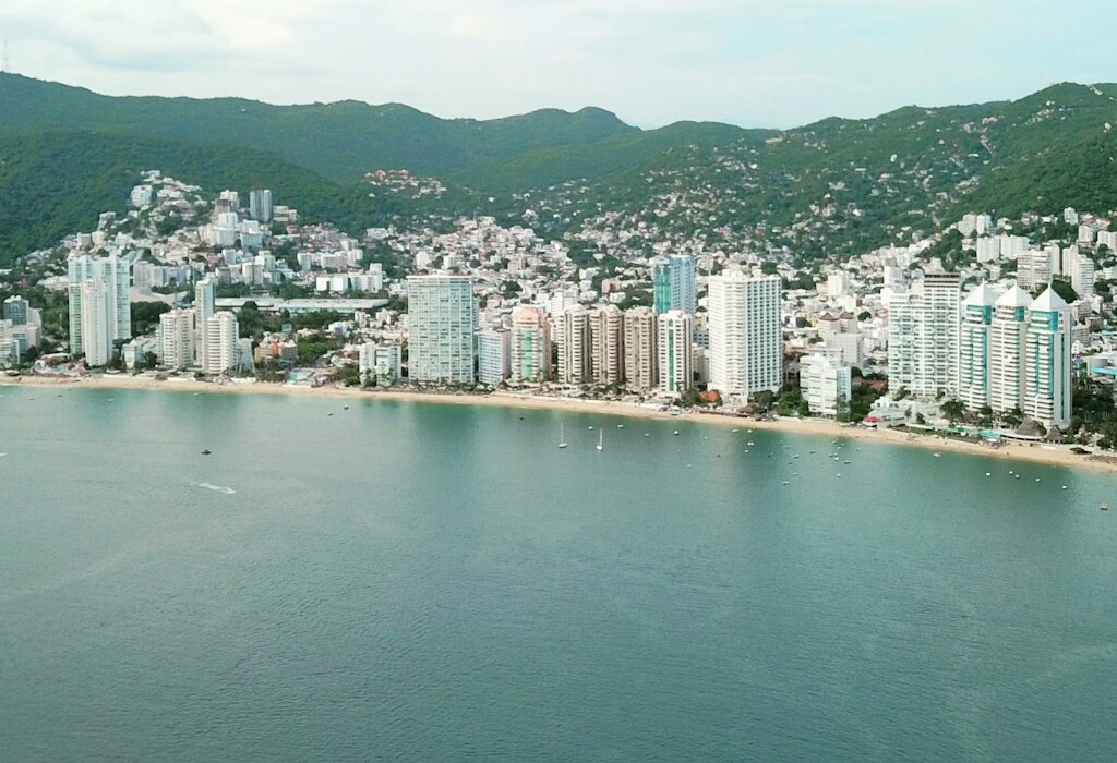 A panoramic view of the Acapulco beach and city skyline, with lush green hills surrounding the coastline and tall buildings by the shore.