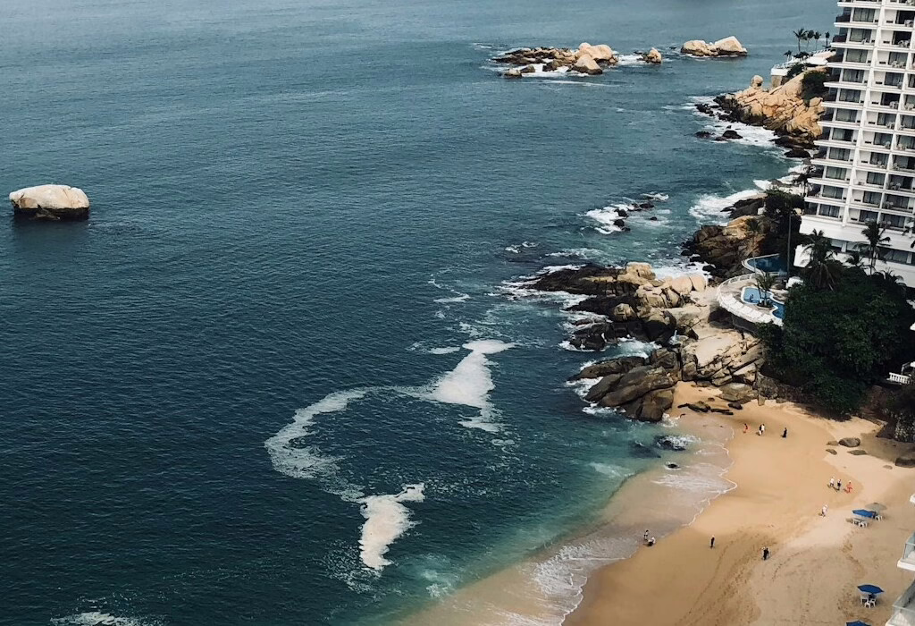 The rugged coastline of Acapulco with waves crashing against the rocks and a sandy beach in the background, offering a scenic ocean view.