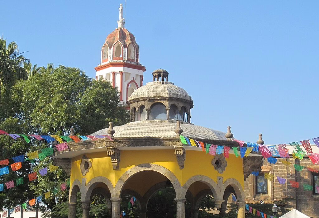 Gazebo colorido e torre de igreja em Tlaquepaque, Jalisco, cercados por papel picado e atmosfera festiva.
