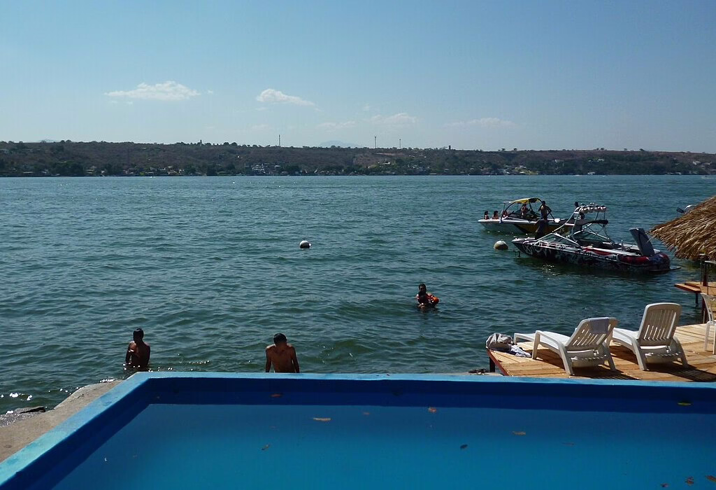 Tourists enjoying a sunny day at Playa La Virgen in Lake Tequesquitengo, featuring a swimming pool, people in the lake, and motorboats anchored near the shore.