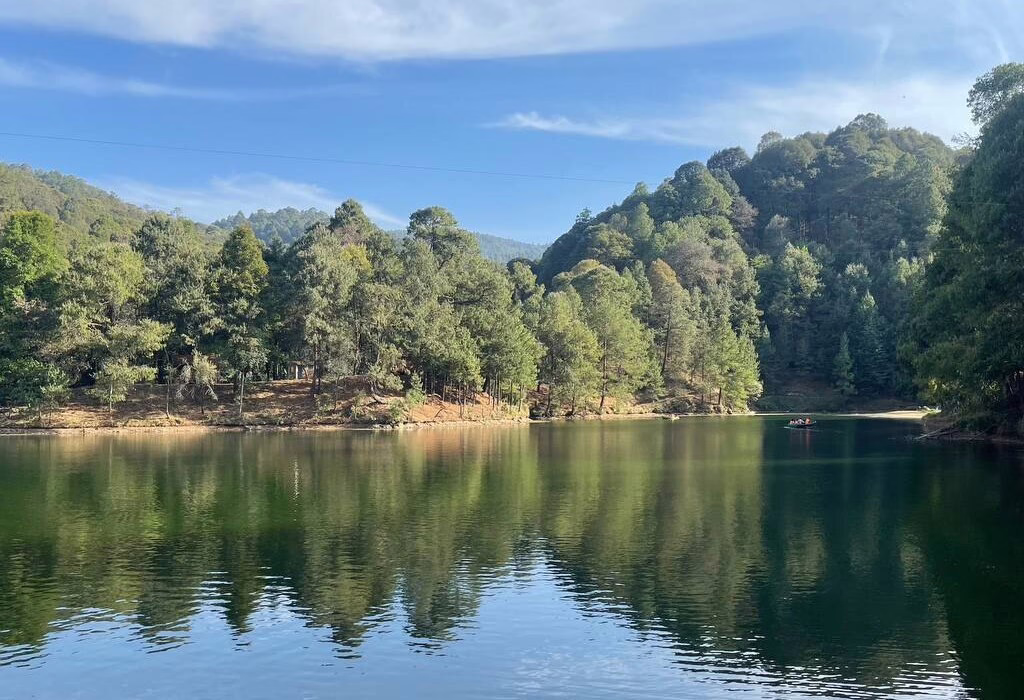 Panoramic view of Presa del Llano in Villa del Carbón on a sunny day, featuring emerald green water with a small boat and pine trees reflecting clearly on the surface under a blue sky.