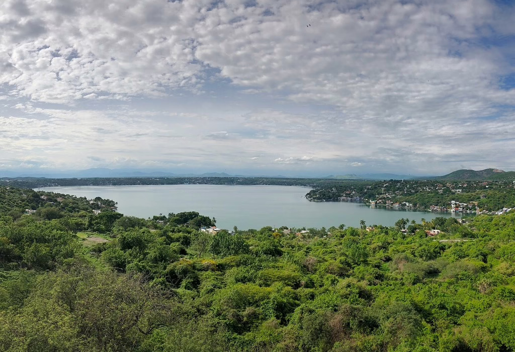 Panoramic view of Lake Tequesquitengo in Morelos, Mexico, surrounded by lush tropical vegetation and houses along the shore under a cloudy blue sky.