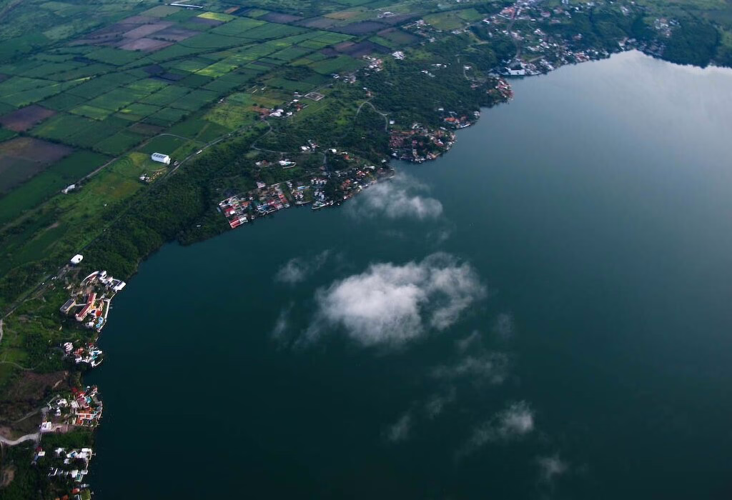 Aerial view of Lake Tequesquitengo in Morelos, Mexico, showing the coastline, luxury houses, and surrounding green fields under a light morning mist.
