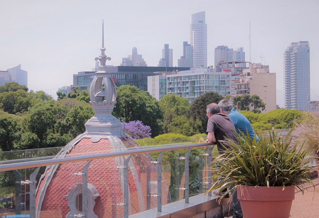 A high angle view from a rooftop terrace looking out over trees, a red-tiled cupola, and modern skyscrapers in Mexico City. Two people lean on a glass railing.
