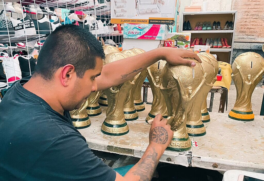 A man with arm tattoos is sitting at a workbench painting a golden World Cup trophy replica in a busy outdoor market stall in Mexico City.