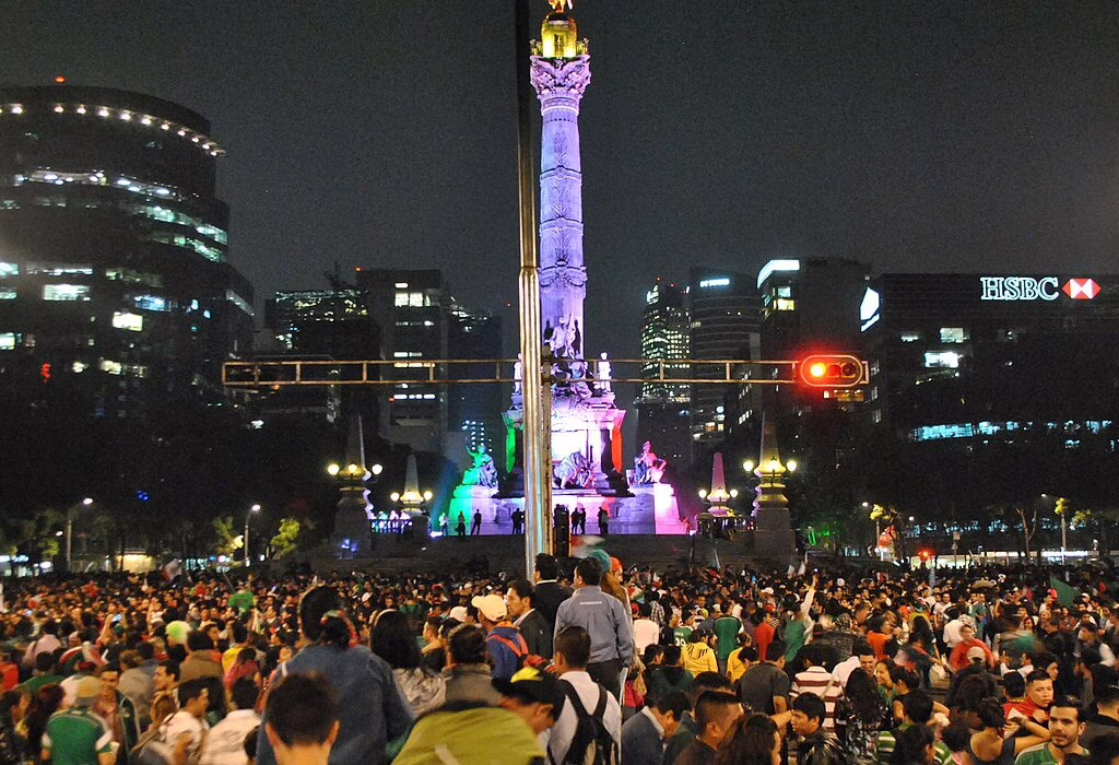 A massive crowd of people gathered around the Angel of Independence monument at night in Mexico City. Football fans celebrating after a World Cup match.