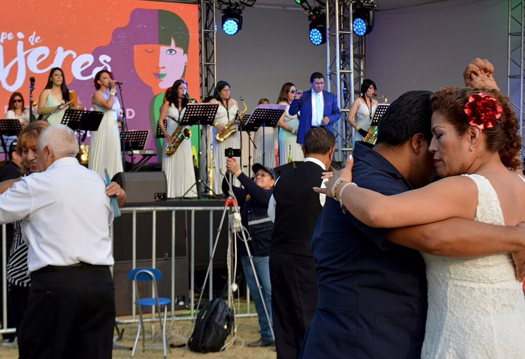 Close-up view of couples dancing closely on a street while an all-female orchestra with saxophones plays live music on a stage with a "Mujeres" banner.