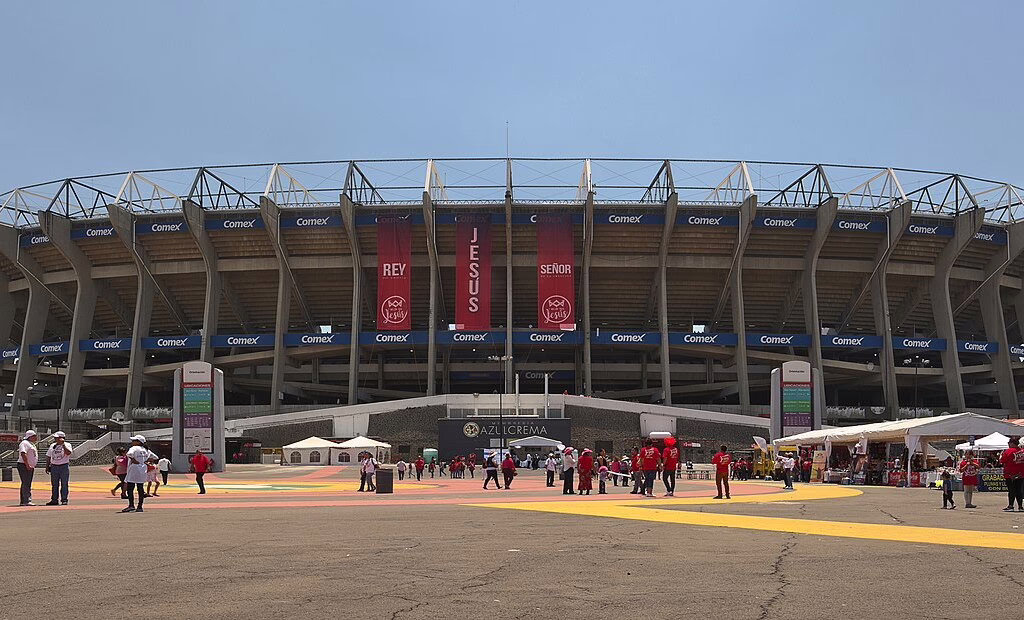 Wide shot of the Estadio Azteca east stands facade with Comex and Telcel banners. Fans in red and white shirts walk on the main plaza toward the entrance, showing event stalls under a clear blue sky.