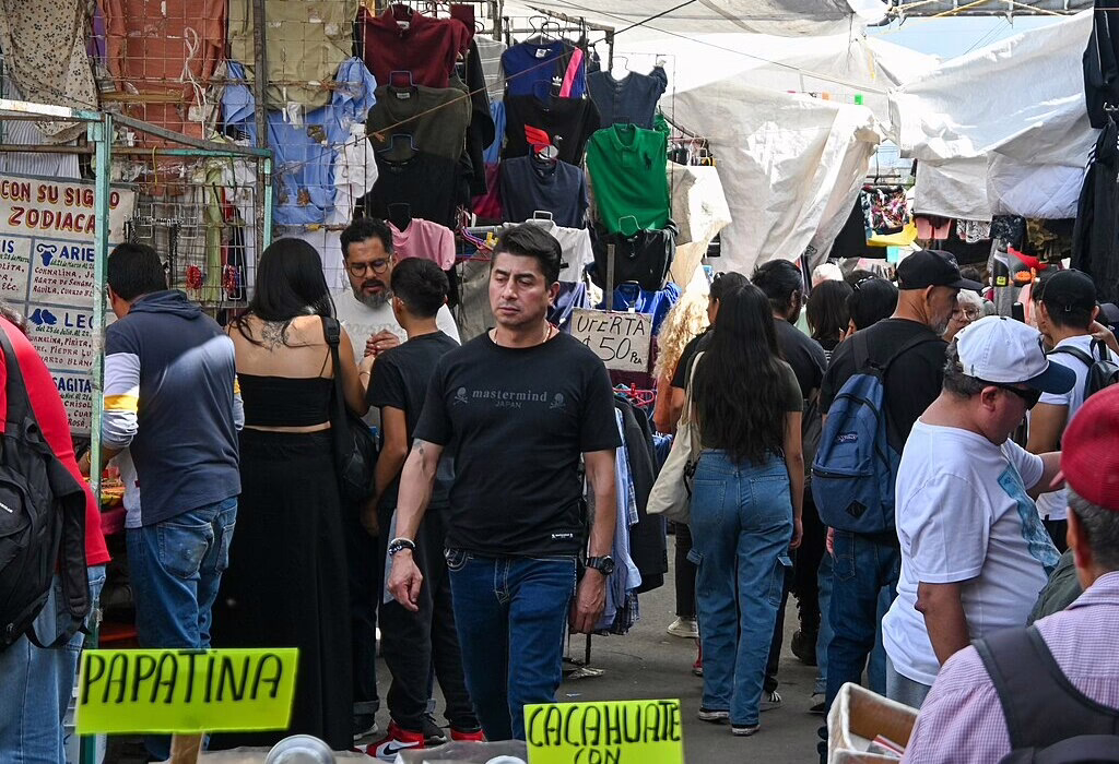 A crowded outdoor street market in Mexico City called La Lagunilla with people walking past stalls selling t-shirts and clothing with hand-written signs.