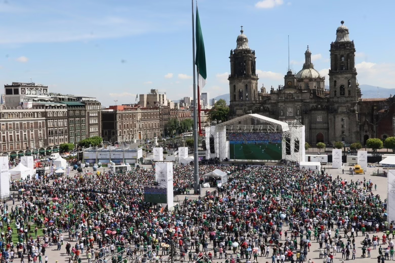 Panoramic view of thousands of people watching a football match at the Zocalo Fan Festival in Mexico City with the Metropolitan Cathedral in the background.