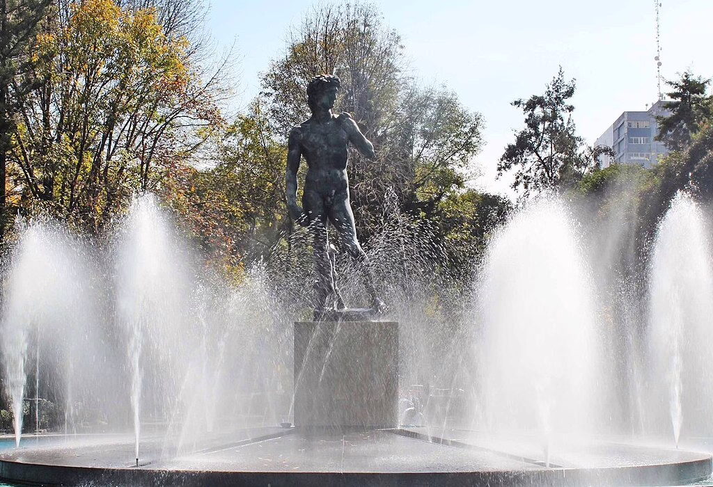 A dark statue of a man standing in the middle of a big water fountain. There are many trees in the background and bright sunlight.
