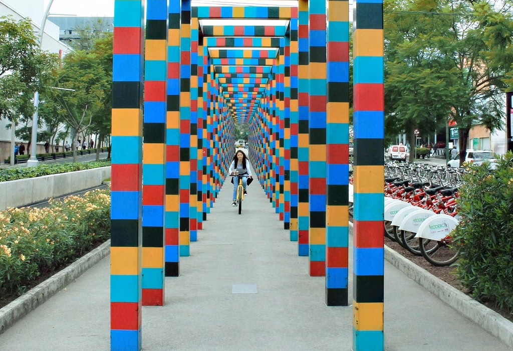 A girl riding a yellow bicycle through many colorful arches on a path. There are many white and red bikes parked on the side.