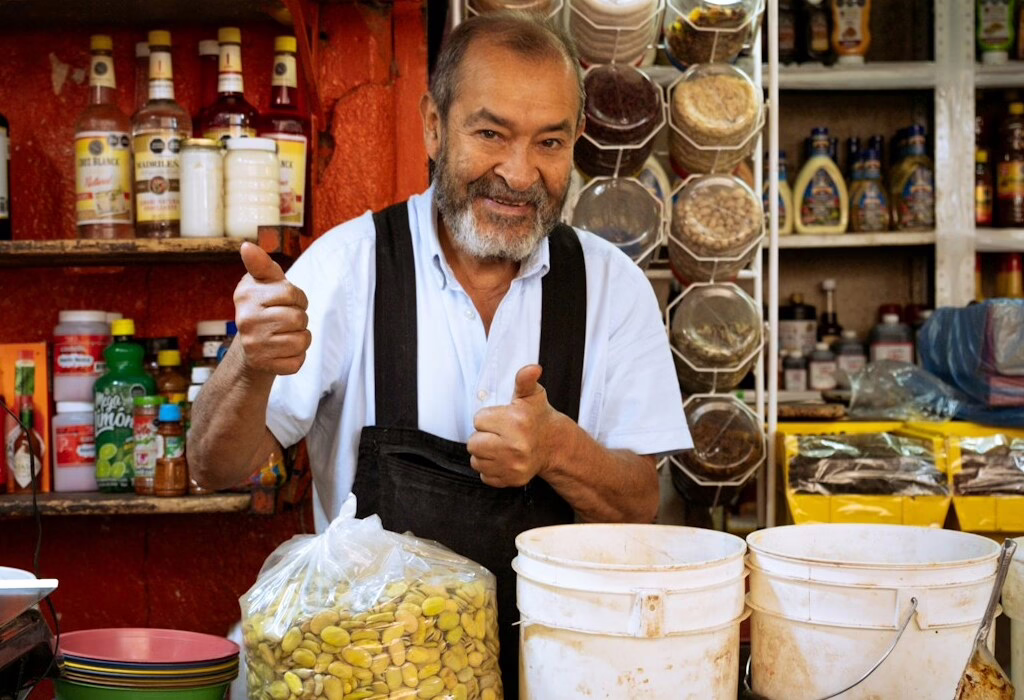 A friendly local man with a beard wearing a black apron giving a thumbs up in his market stall filled with seeds and grains in Mexico City.