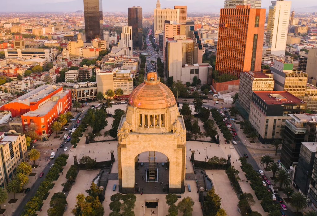 An aerial view of a large stone monument with a copper dome in a big square. There are many tall buildings and streets around it during sunset.