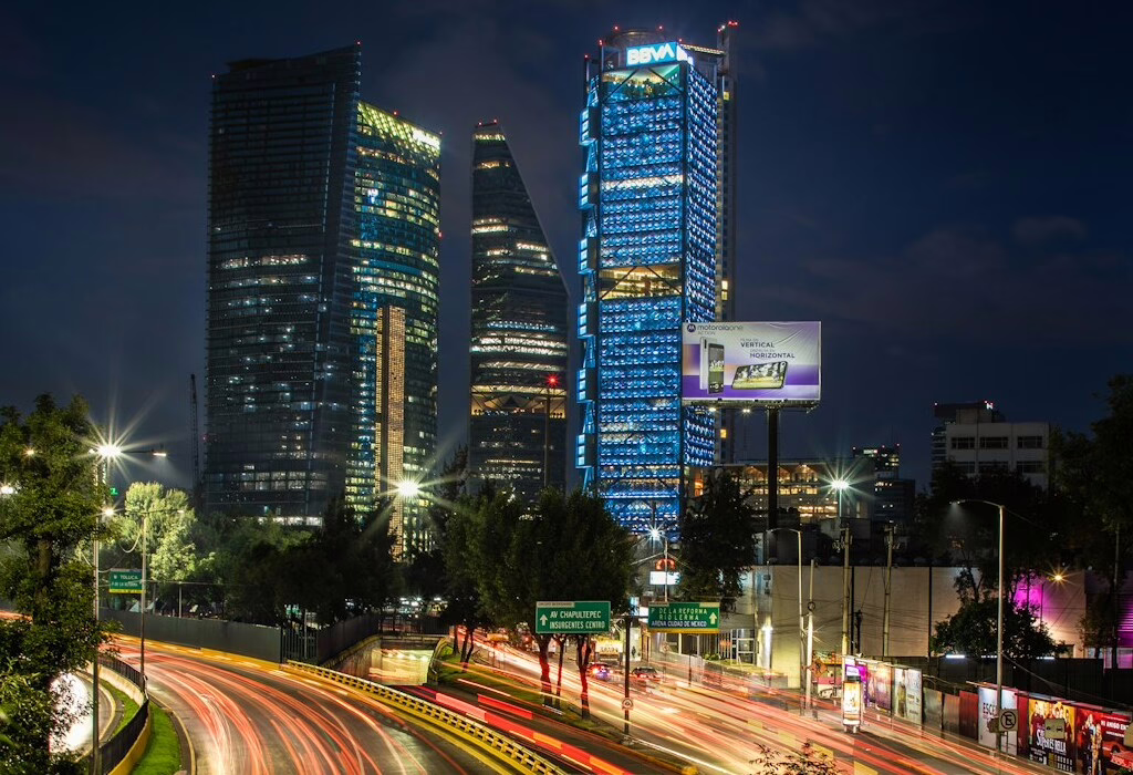 Elevated night shot of the Paseo de la Reforma skyline in Mexico City showing the Torre BBVA Mexico and Torre Reforma. Includes the Motorola One billboard and light trails from traffic near the Chapultepec tunnel, a primary area for fans during FIFA World Cup 2026.
