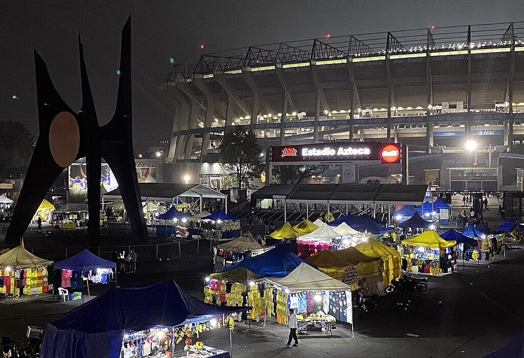An aerial night view of the illuminated Estadio Azteca in Mexico City with many colorful market stalls and food stands in the foreground.